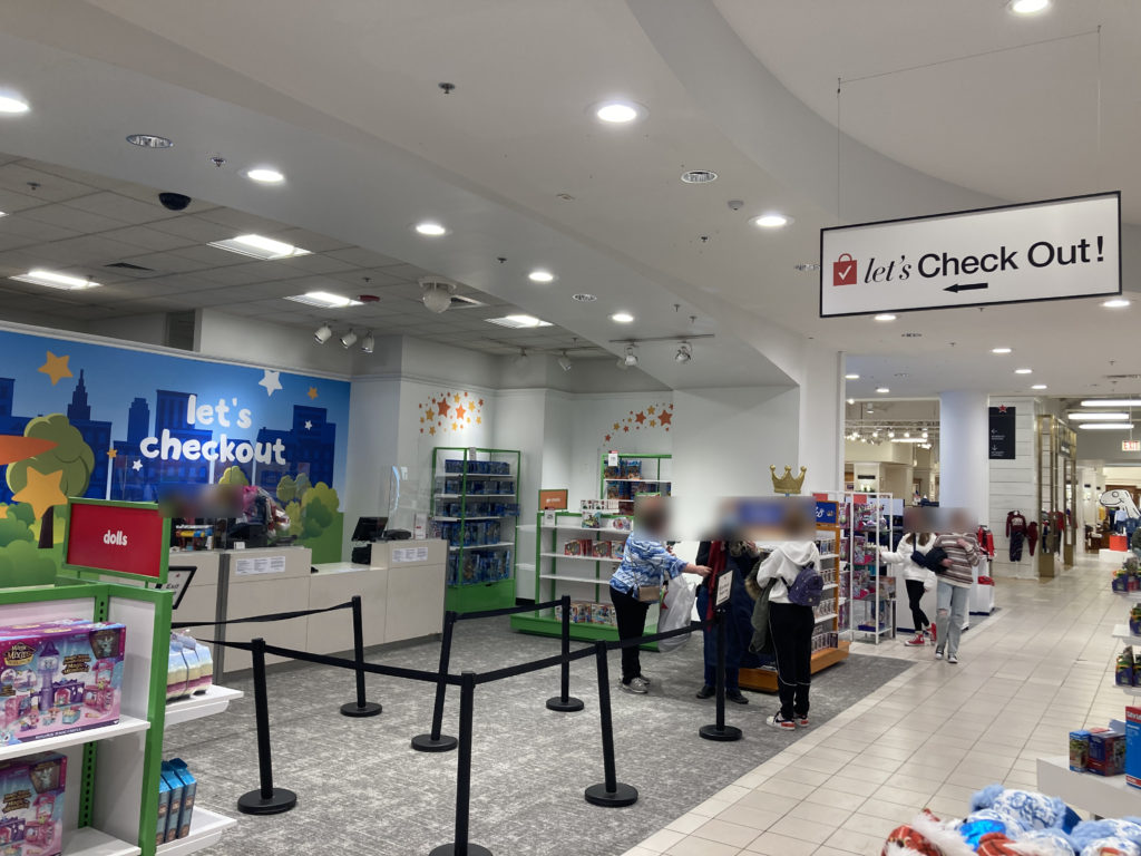 A photo of a checkout area. On the left is a mural painted in Toys R Us style that says "Let's Checkout". On the right there is a sign hanging that says "Let's Check Out!" using Macy's branding.