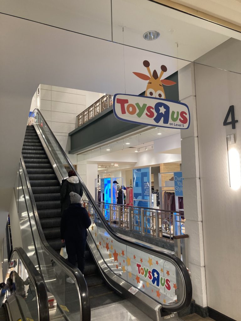 A photo of an escalator in Macy's Chicago Store. A sign hanging above the escalator says "Toys R Us on Level 5".