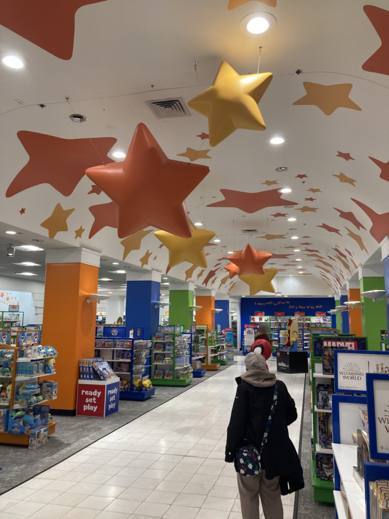 An aisleway in a Macy's story. On each side of the aisle there are shelves with toys, and there are large orange and yellow stars painted on the ceiling.