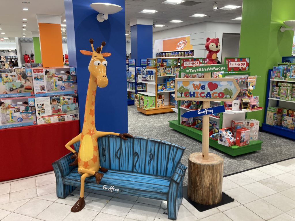 A photo of a small plastic couch, on which Geoffrey the Giraffe sits.